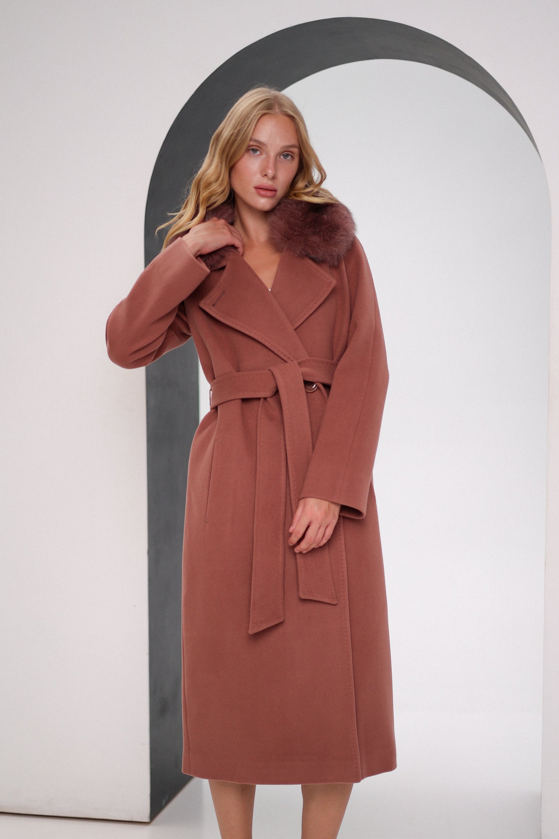 Woman wearing a brown coat with fur collar standing in front of an archway.