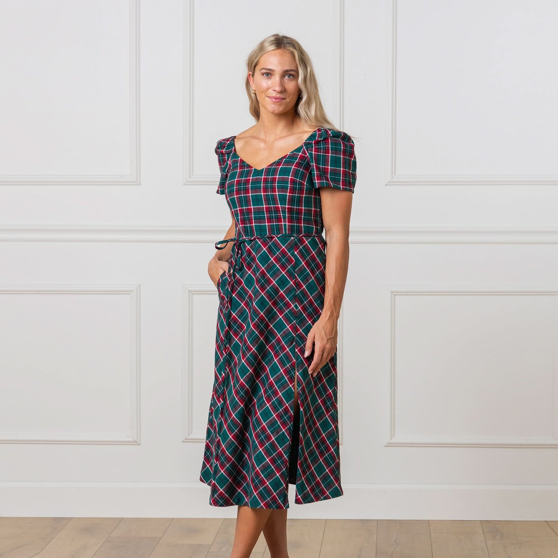 Woman wearing a green and red plaid dress against a white paneled wall.