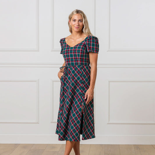 Woman wearing a green and red plaid dress against a white paneled wall.