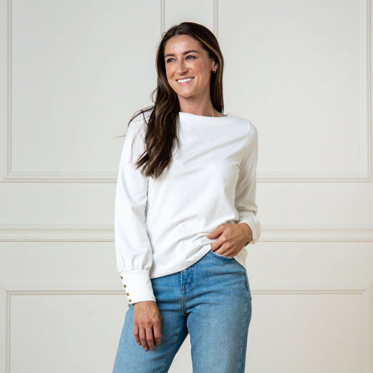 Woman wearing a white sweater and blue jeans against a white paneled wall.