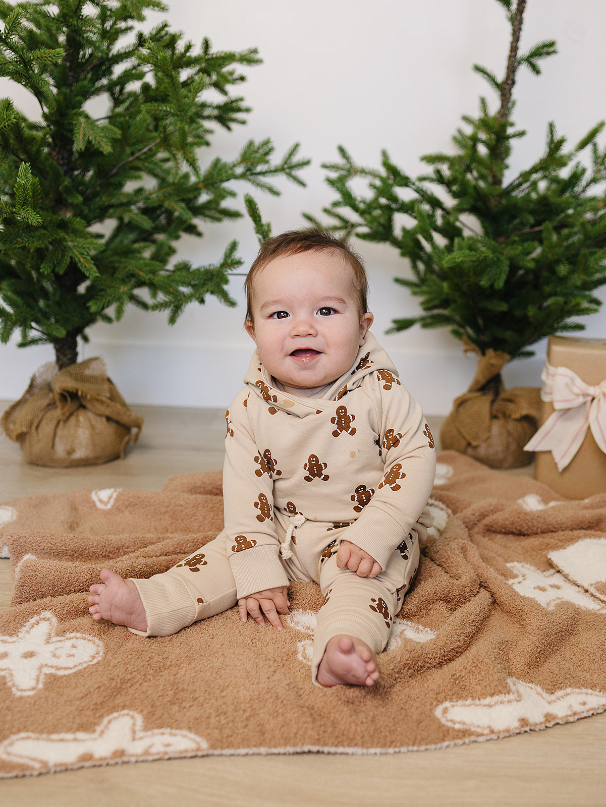 Baby in gingerbread-themed outfit sitting on a blanket with Christmas trees in the background