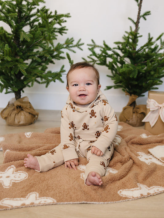 Baby in gingerbread-themed outfit sitting on a blanket with Christmas trees in the background