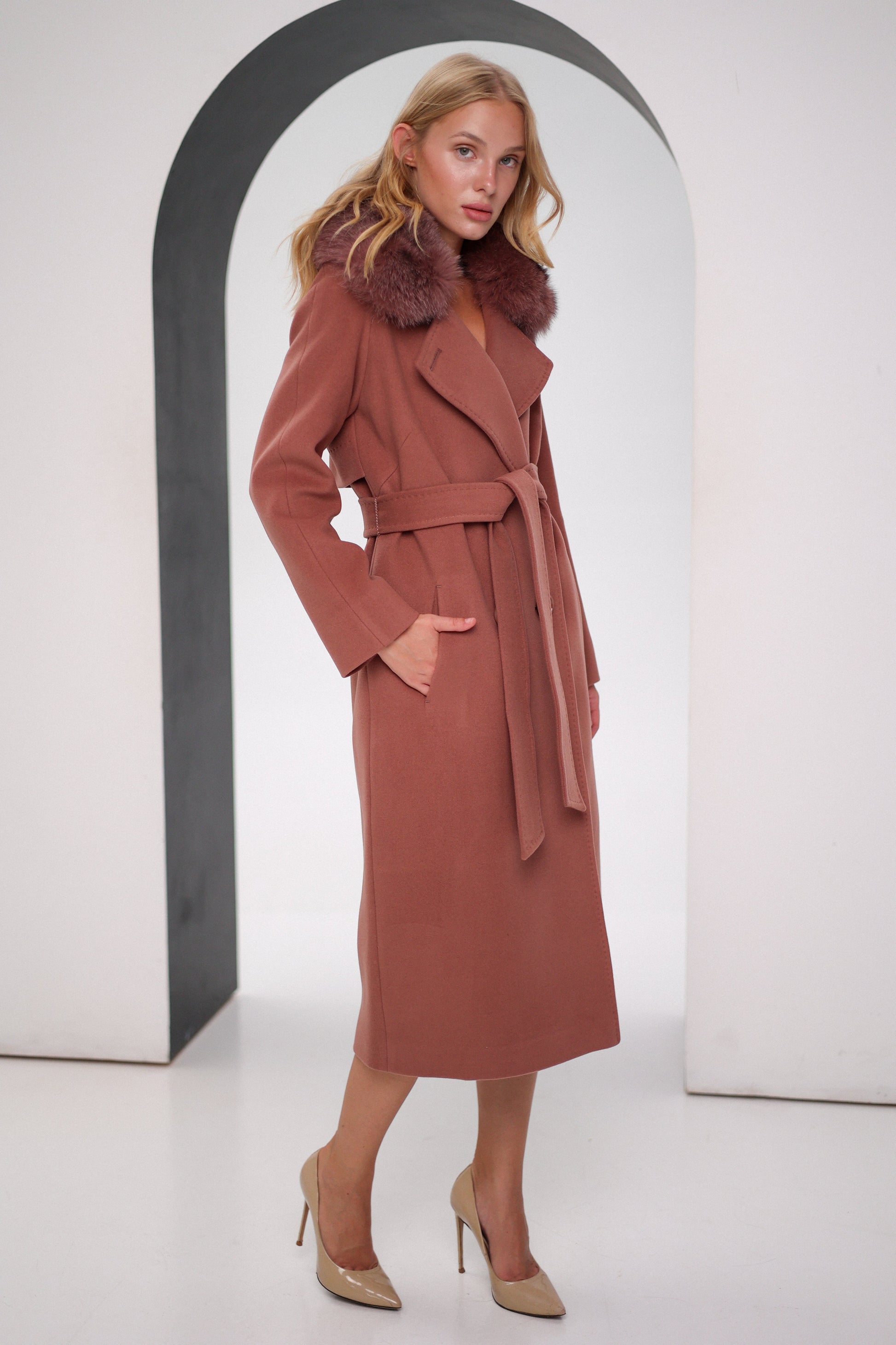 Woman wearing a long brown coat with fur collar standing in front of an archway.