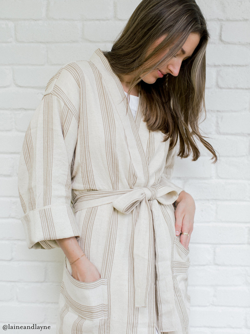Woman wearing a beige striped robe against a white brick wall.