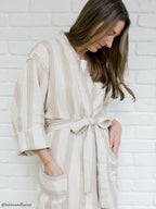Woman wearing a beige striped robe against a white brick wall.