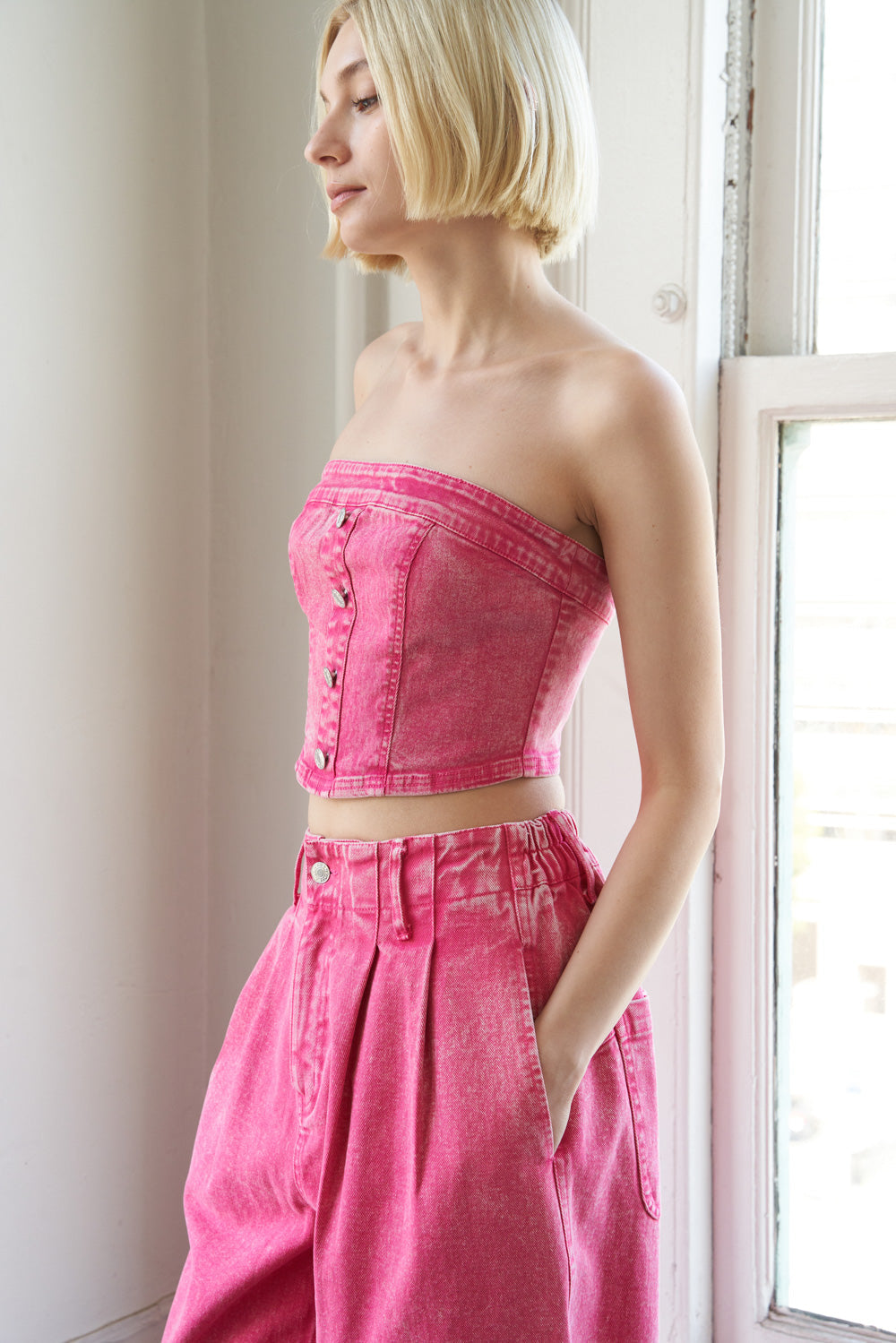 Woman wearing a pink strapless top and high-waisted pants standing by a window.