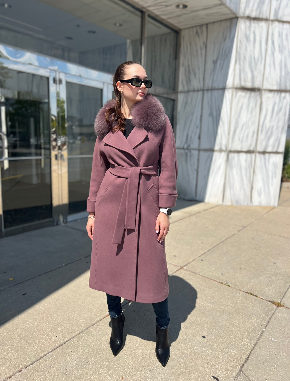 Woman wearing a long pink coat with a fur collar standing outside a building.