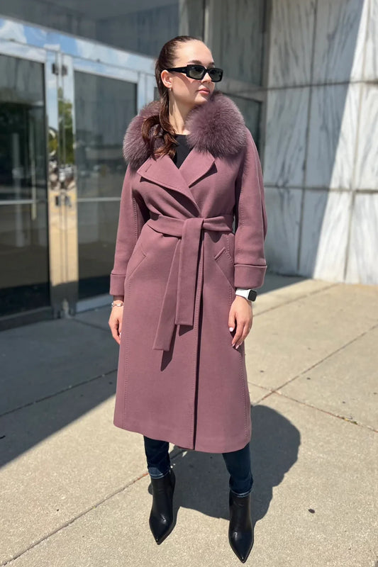 Woman wearing a long pink coat with fur collar standing on a sidewalk.