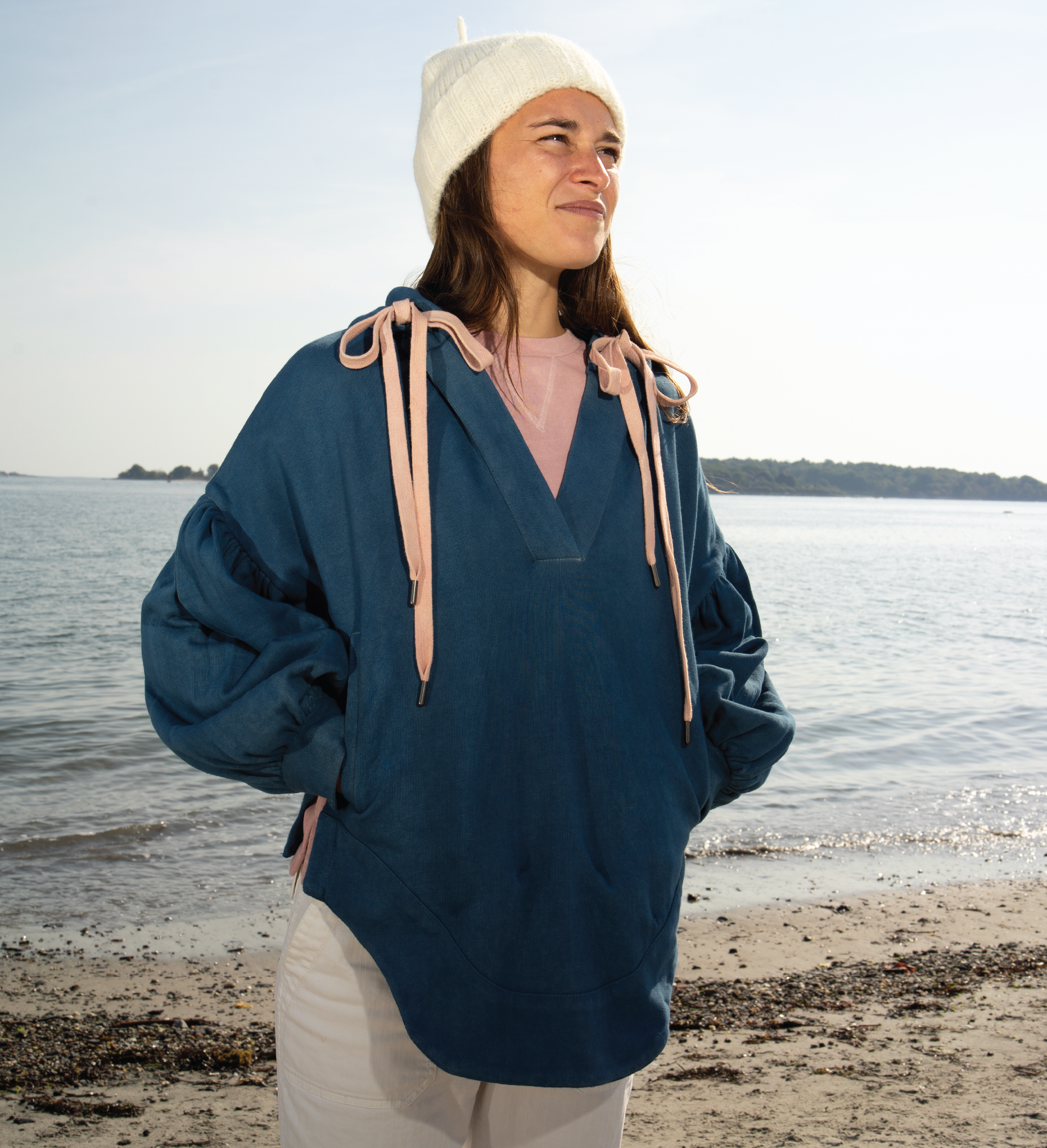 Person wearing a blue hoodie and beige beanie standing on a beach with water in the background