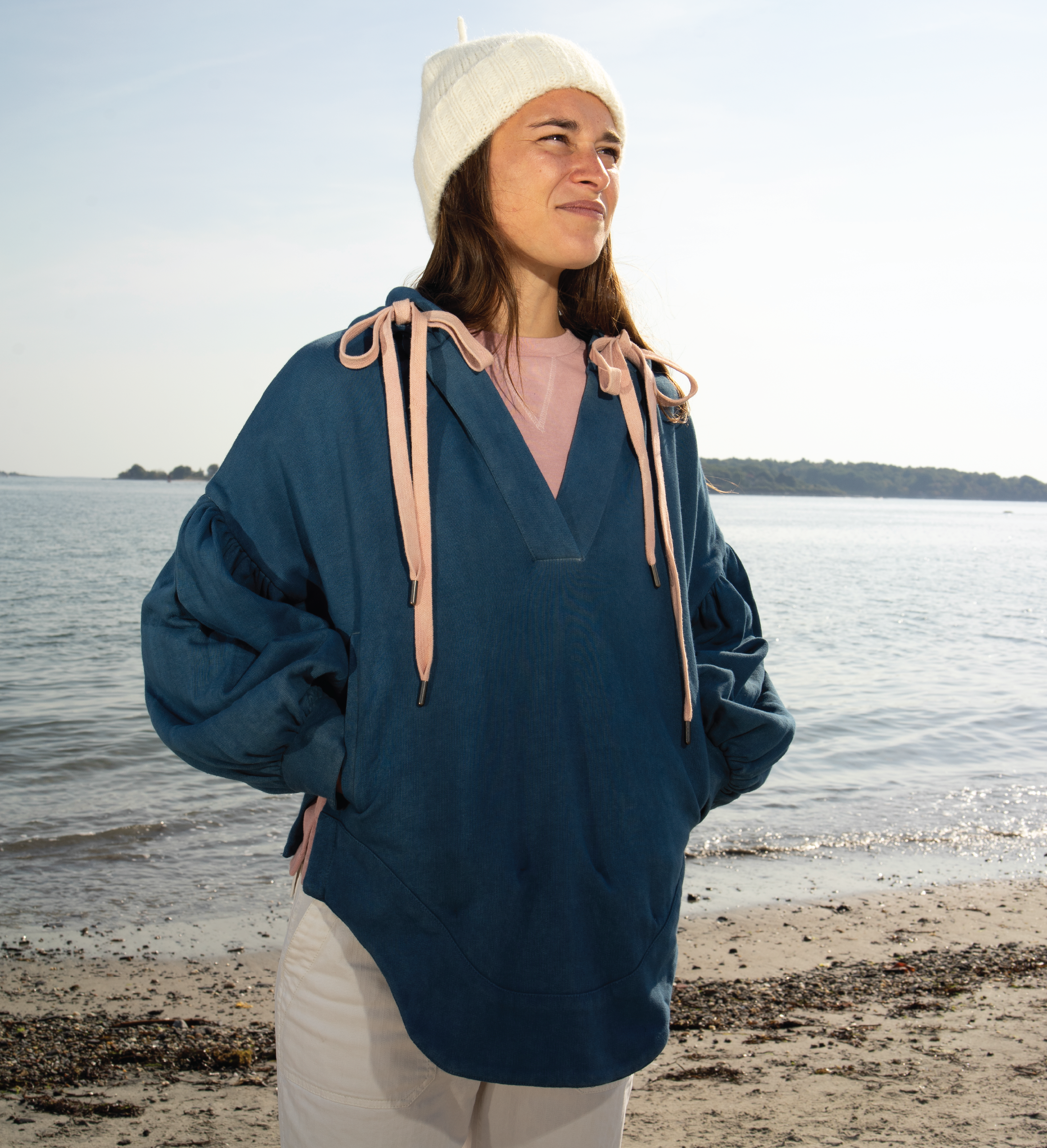 Person wearing a blue hoodie and beige beanie standing on a beach with water in the background