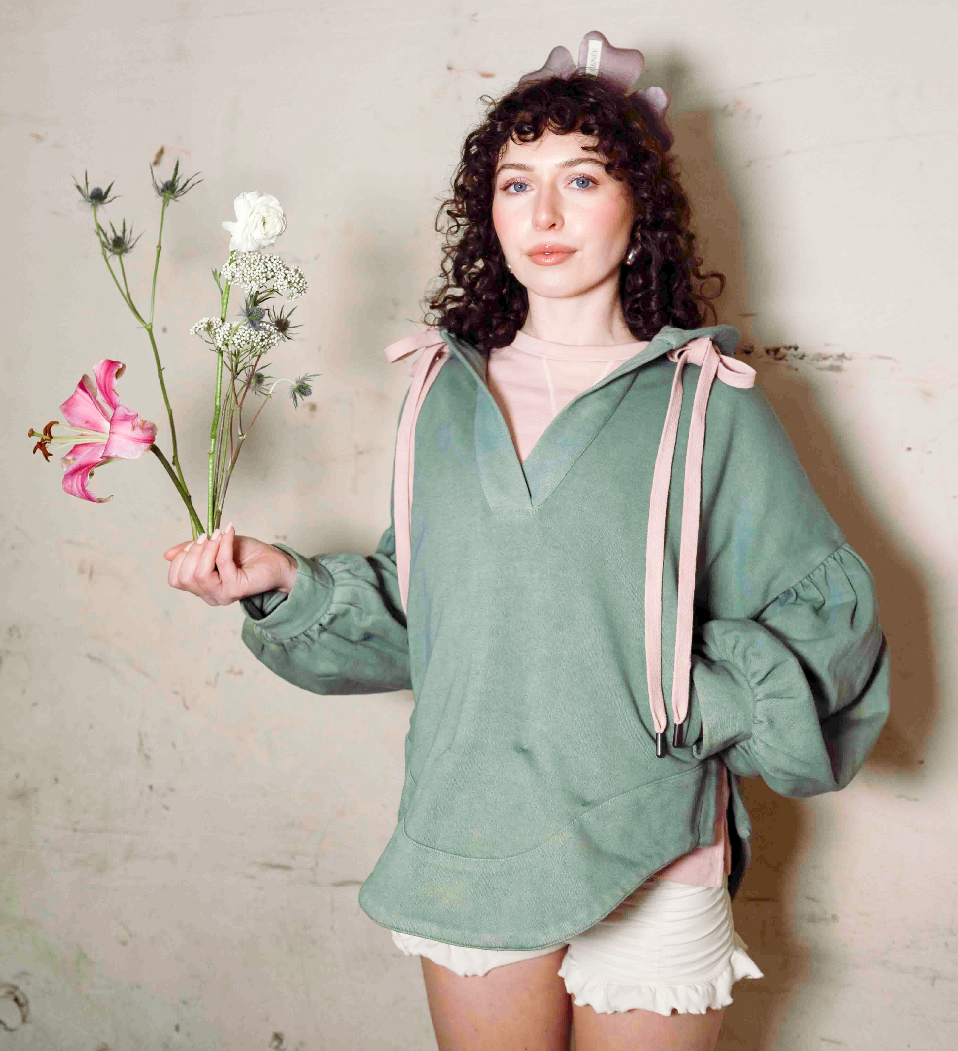 Woman wearing a green hoodie with pink ribbons, holding flowers against a textured wall.