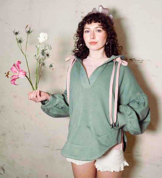 Woman wearing a green hoodie with pink ribbons, holding flowers against a textured wall.