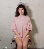 Person wearing a pink oversized shirt sitting on a chair against a textured wall.