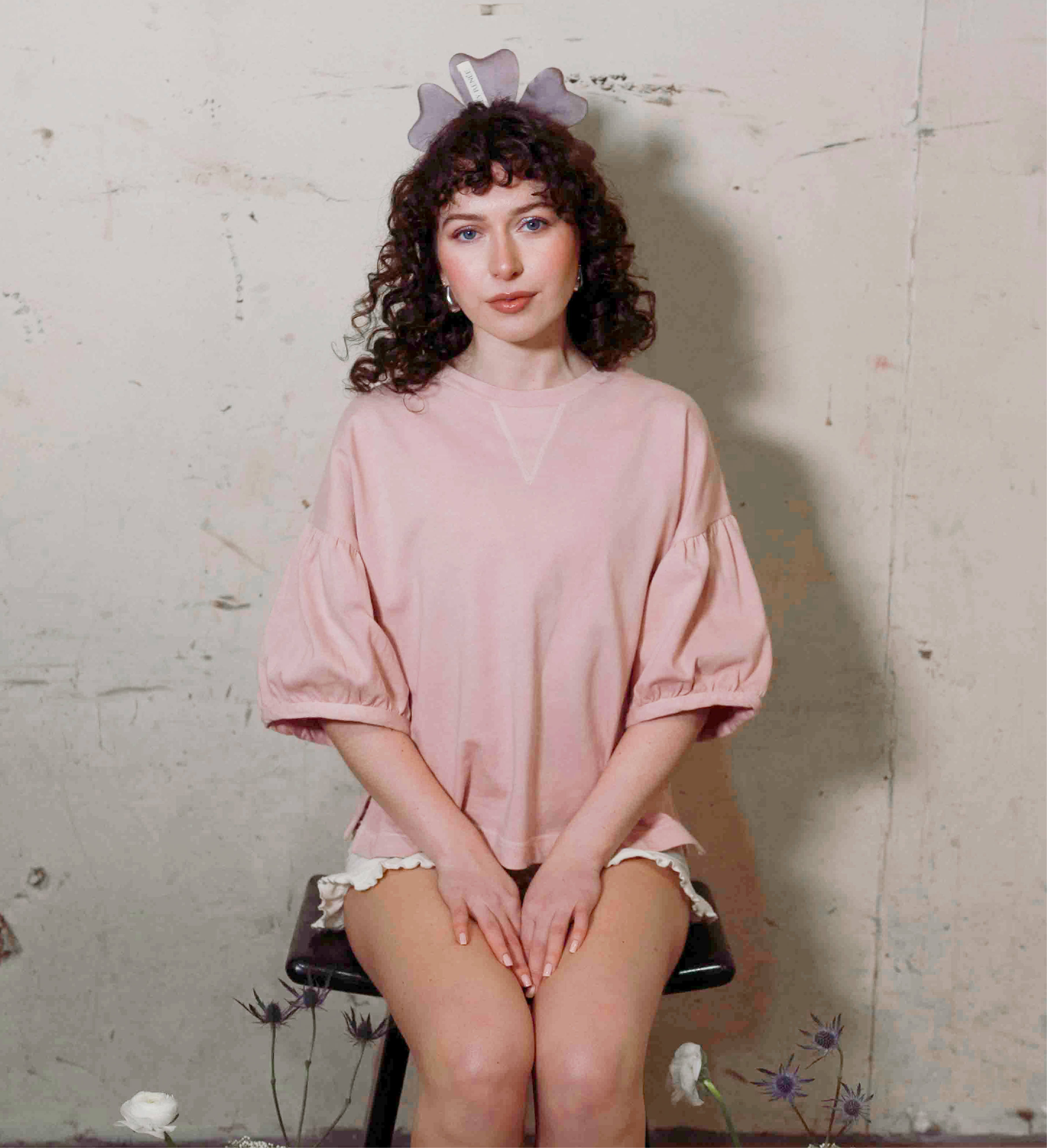 Person wearing a pink oversized shirt sitting on a chair against a textured wall.