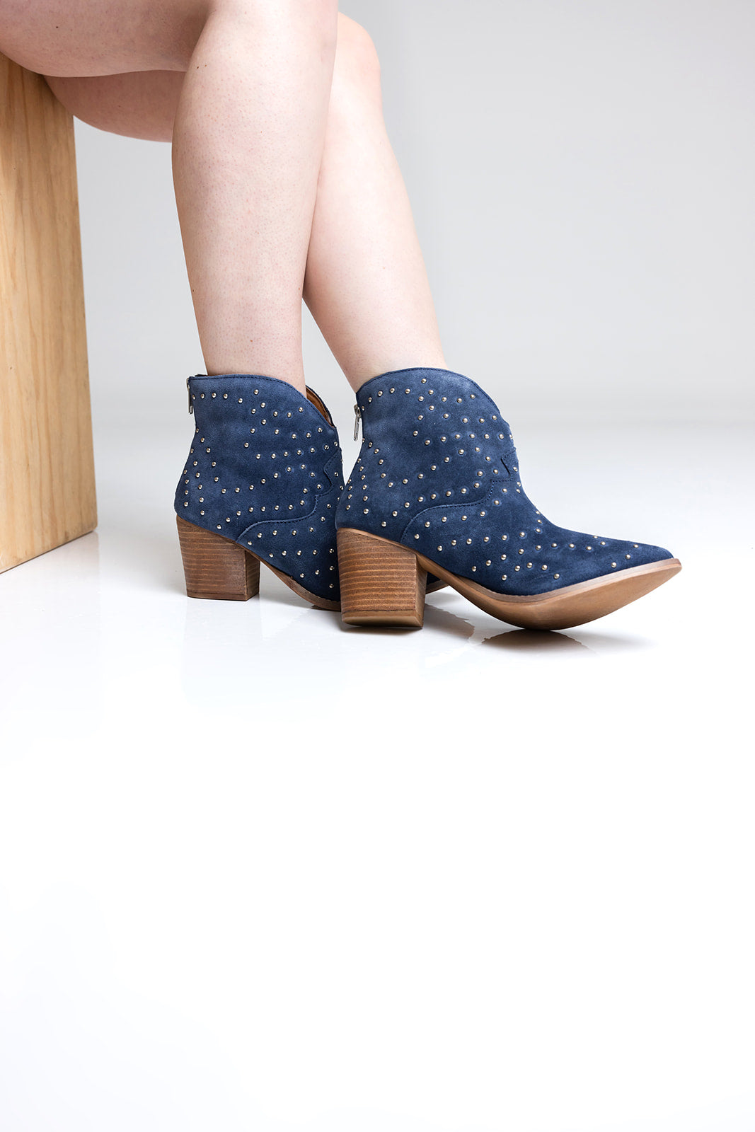 Navy blue studded ankle boots with brown soles worn by a person sitting on a wooden stool.