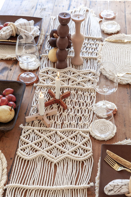 Decorative table setting with macrame runner, candles, and glasses on a wooden table.