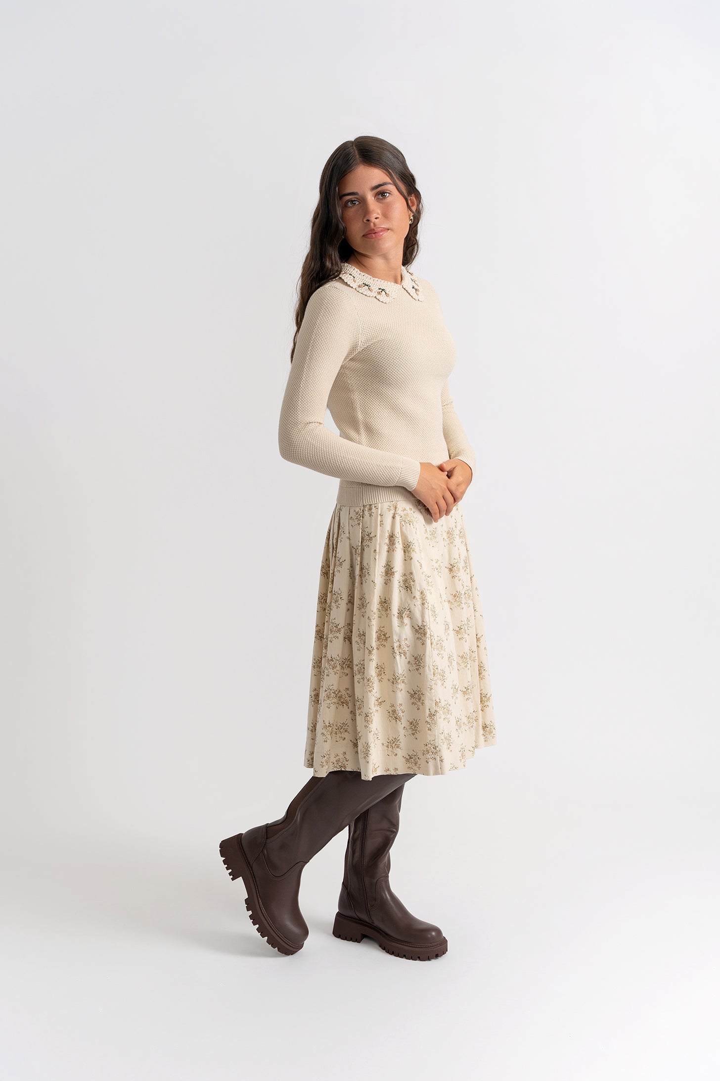 Woman wearing a beige sweater and patterned skirt with brown boots on a white background