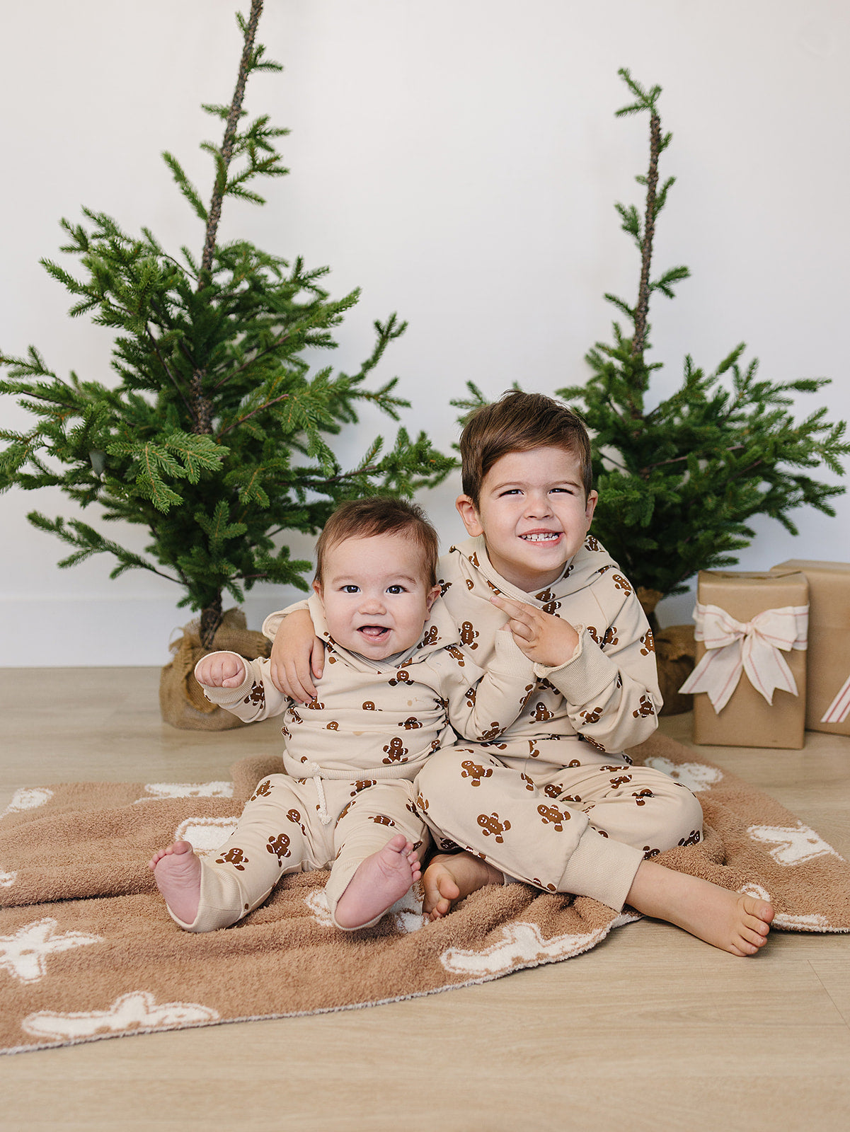 Two children in matching pajamas sitting on a blanket with Christmas trees and presents in the background.