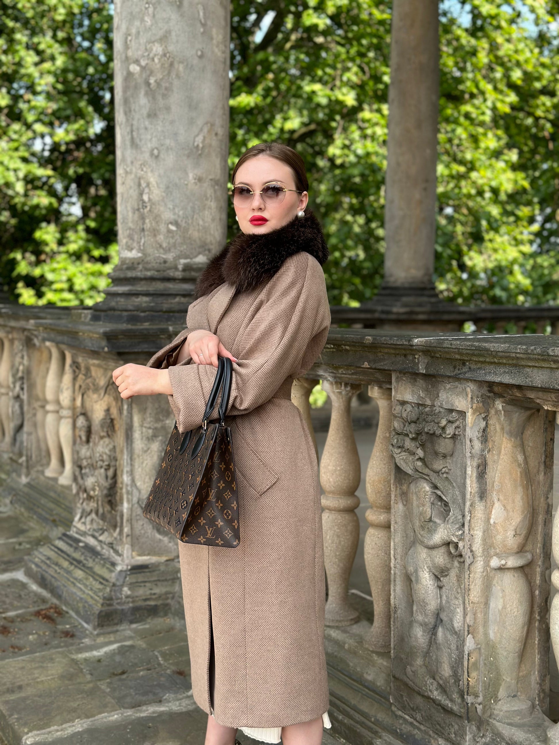 Woman in a beige coat standing on a stone balcony with greenery in the background