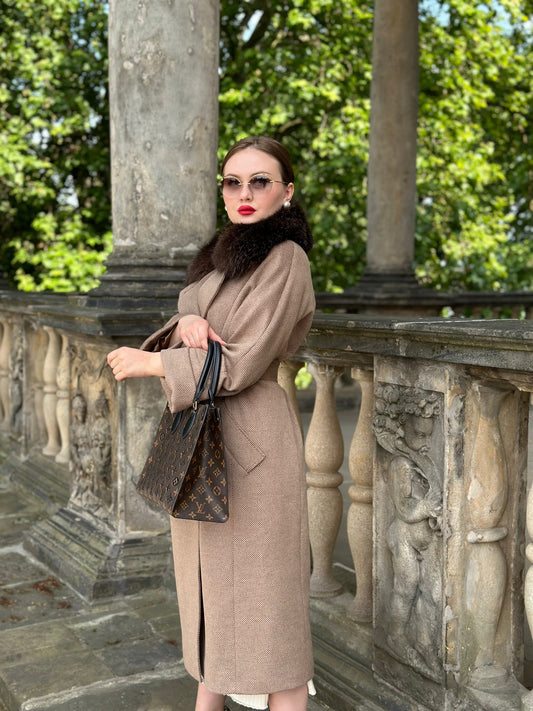Woman in a beige coat standing on a stone balcony with greenery in the background