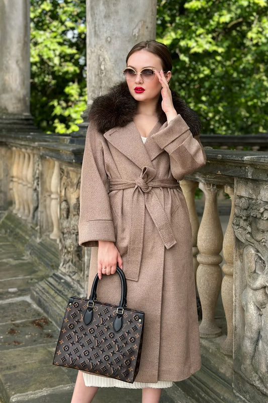 Woman in a beige coat with fur collar holding a textured handbag outdoors.